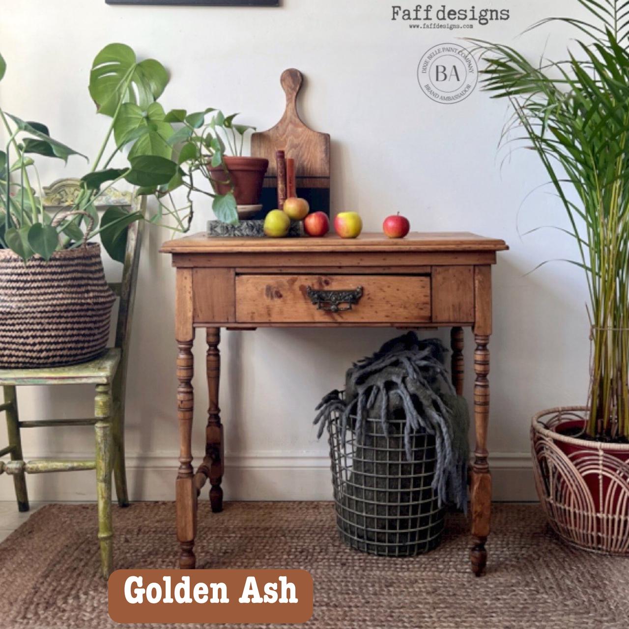 A rustic wooden table holds fruits, cutting boards, and a candle. It's flanked by green plants in woven baskets. A vintage chair and textured rug complete the cozy setting. Text reads "Golden Ash" and "Faff designs.