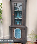 A vintage corner cabinet with a gray finish and blue interior, featuring geometric glass panels. It's decorated with potted plants on either side. The text "Up In Smoke" is visible at the bottom left.
