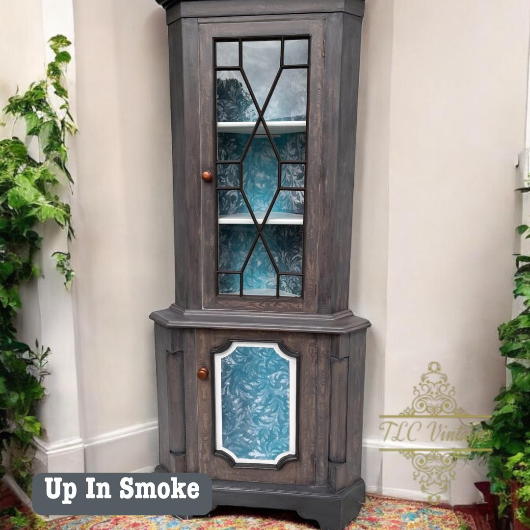 A vintage corner cabinet with a gray finish and blue interior, featuring geometric glass panels. It's decorated with potted plants on either side. The text "Up In Smoke" is visible at the bottom left.