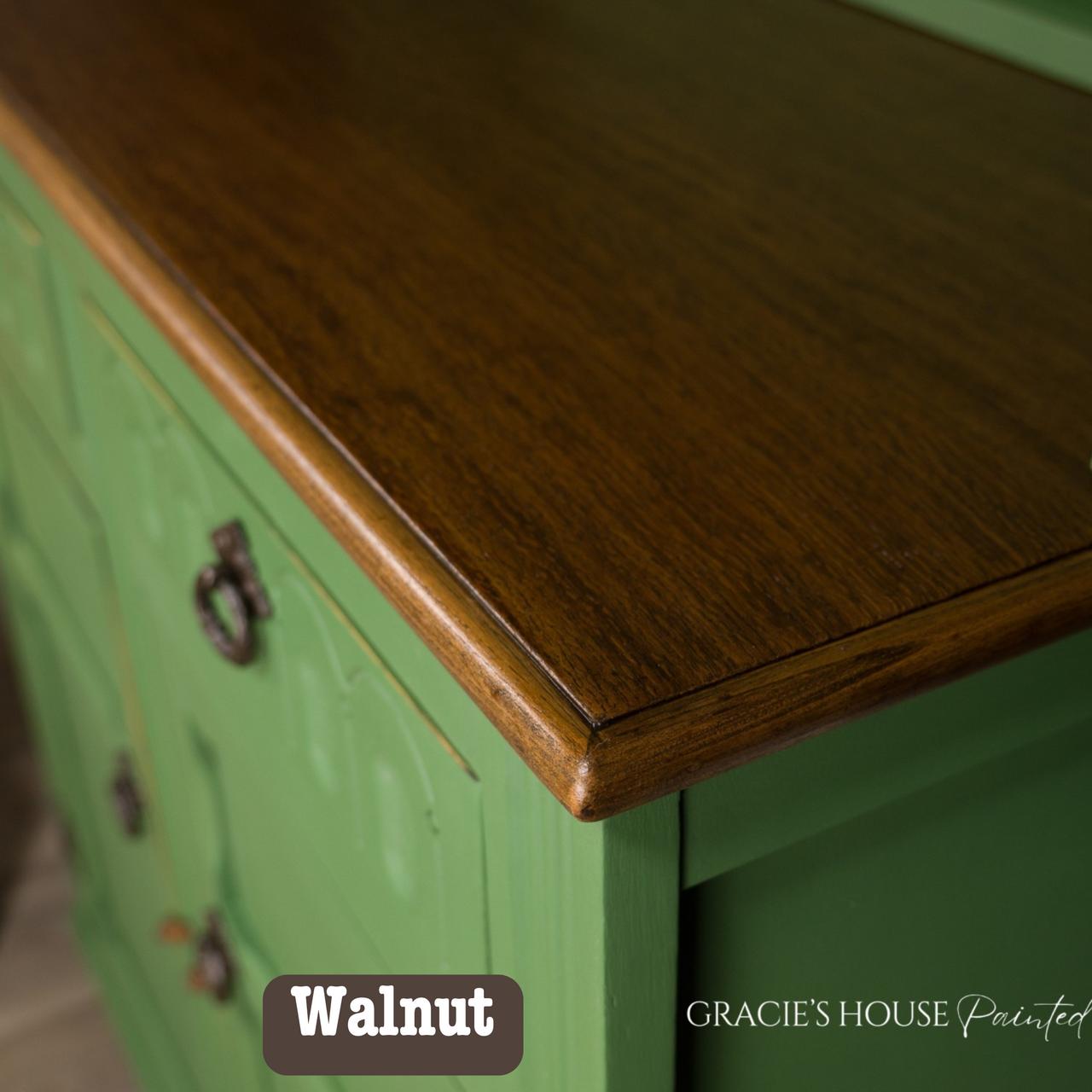 Close-up of a wooden cabinet with a green painted base and a dark walnut-stained top. The surface is glossy, showing a smooth texture. Three decorative metal handles are visible. The words "Walnut" and "Gracie's House Painted" appear on the image.
