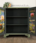 A vintage cabinet with open doors reveals empty shelves. The inner doors feature painted water lilies on a pond. A small potted plant sits on top. The wall behind shows "Gracie's House Painted Furniture." Wooden floor completes the scene.