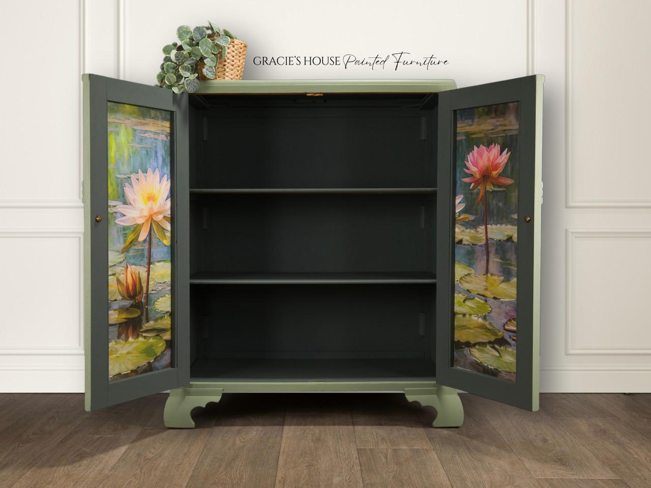 A vintage cabinet with open doors reveals empty shelves. The inner doors feature painted water lilies on a pond. A small potted plant sits on top. The wall behind shows "Gracie's House Painted Furniture." Wooden floor completes the scene.