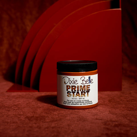 A jar of Dixie Belle Prime Start stands on a red surface with abstract, curved red panels in the background, all under dramatic lighting.