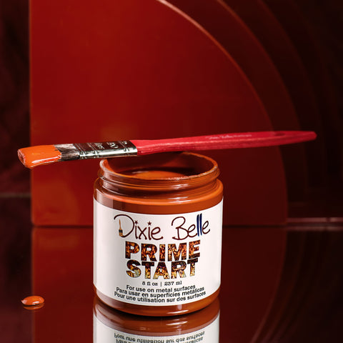A jar of Dixie Belle Prime Start paint with an orange paint-dipped brush resting on top. Some paint drips down the jar, and the background features layered, curved, reddish-brown panels.