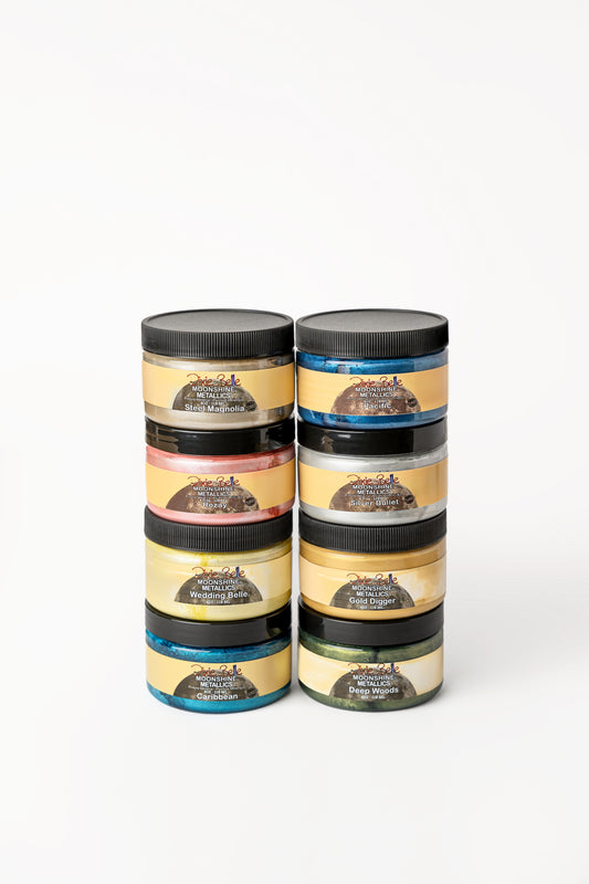Eight jars of Dixie Belle Moonshine Metallics paint are stacked in two columns. Each jar has a black lid and a yellow label, with different colors visible through the containers.