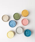 Eight open paint cans arranged on a white surface, each containing a different color: beige, green, yellow, gold, blue, pink, light gray, and teal blue. The cans are viewed from above.