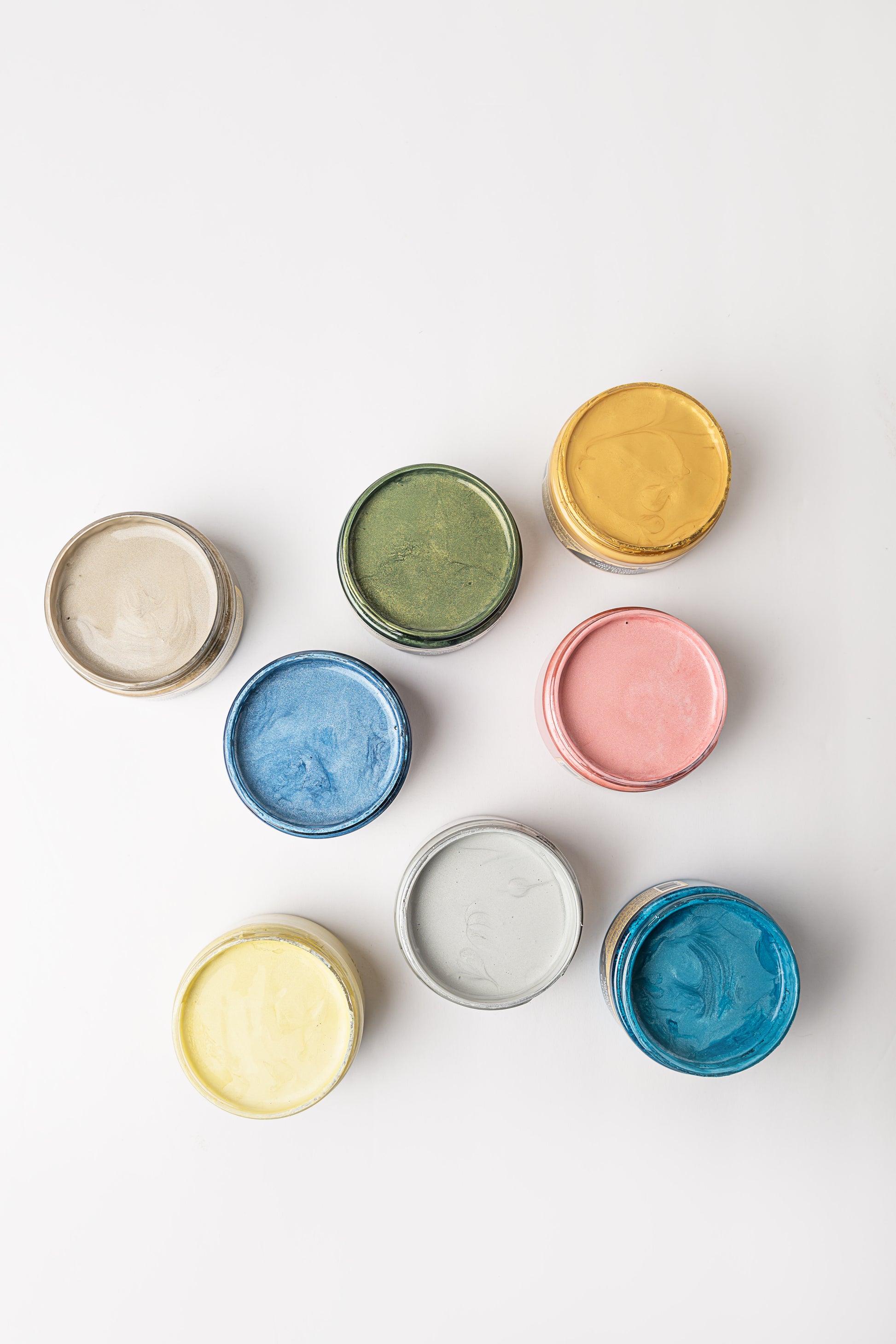 Eight open paint cans arranged on a white surface, each containing a different color: beige, green, yellow, gold, blue, pink, light gray, and teal blue. The cans are viewed from above.