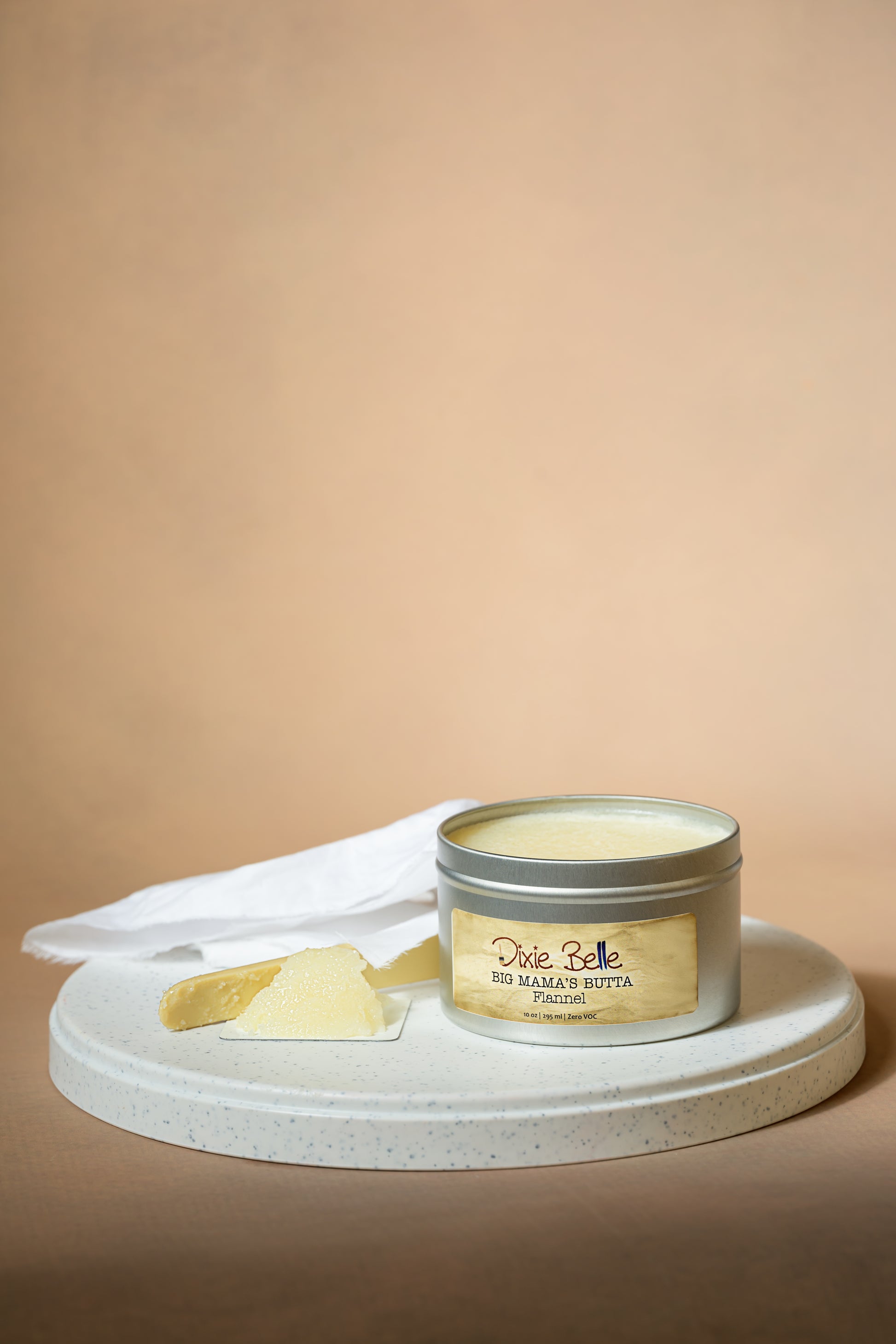 A tin labeled "Dixie Belle Big Mama's Butta Flannel" sits on a round white tray with a creamy solid substance and a piece of white cloth beside it, against a beige background.