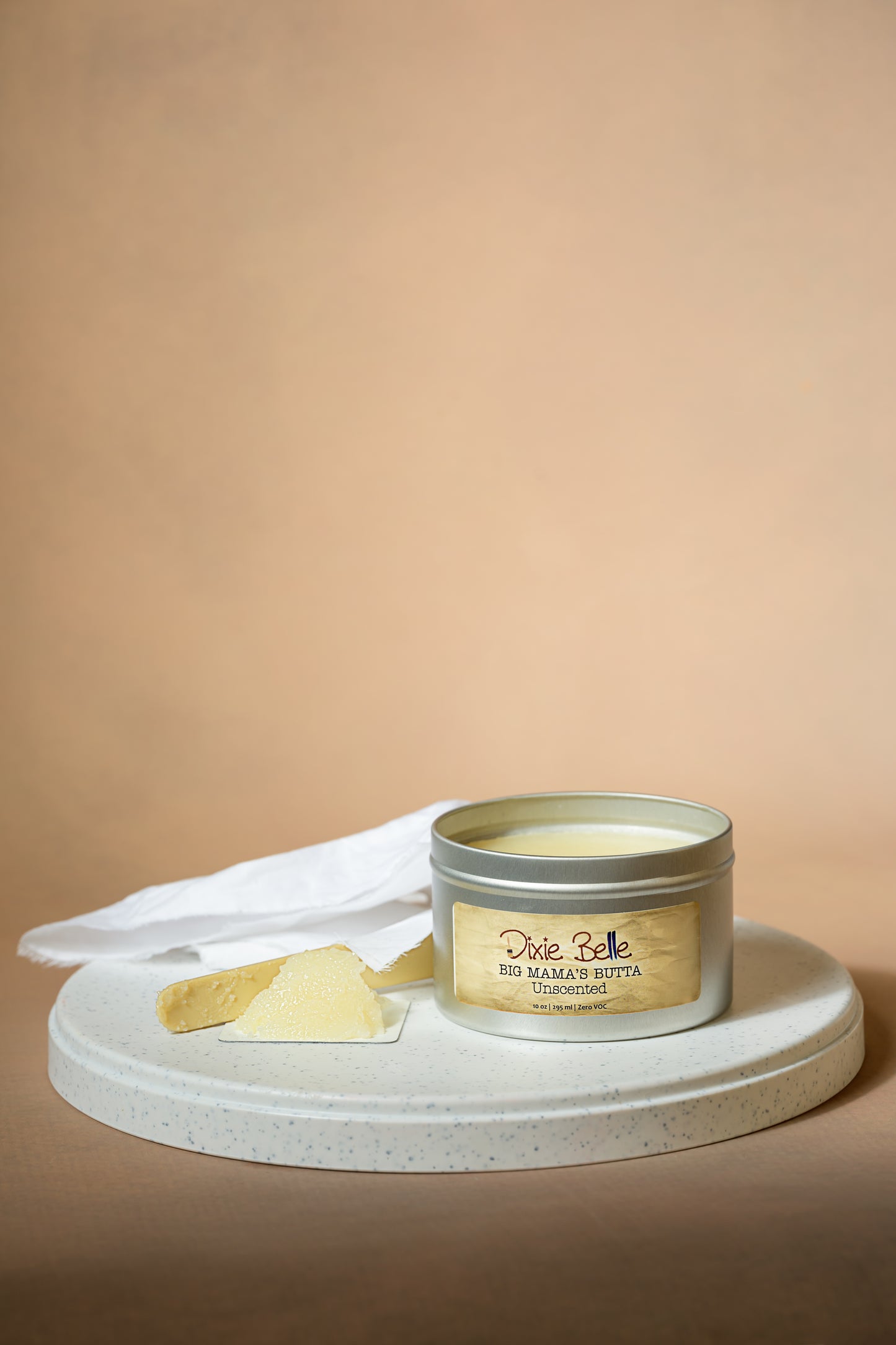 A tin labeled “Dixie Belle Big Mama’s Butta Unscented” sits on a white round tray with a piece of the product and a white cloth beside it, against a beige background.