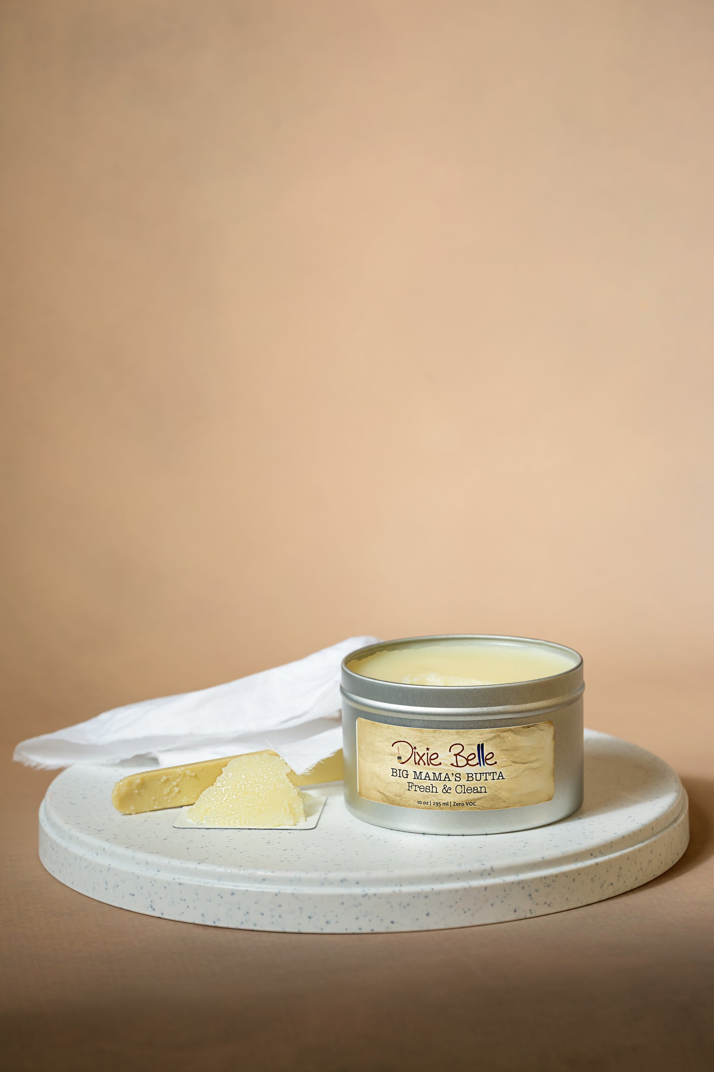 A tin labeled "Dixie Belle Big Mama’s Butta Ginger Zen" sits open on a round white tray, with a cloth and a small portion of the product scooped out beside it, against a neutral beige background.