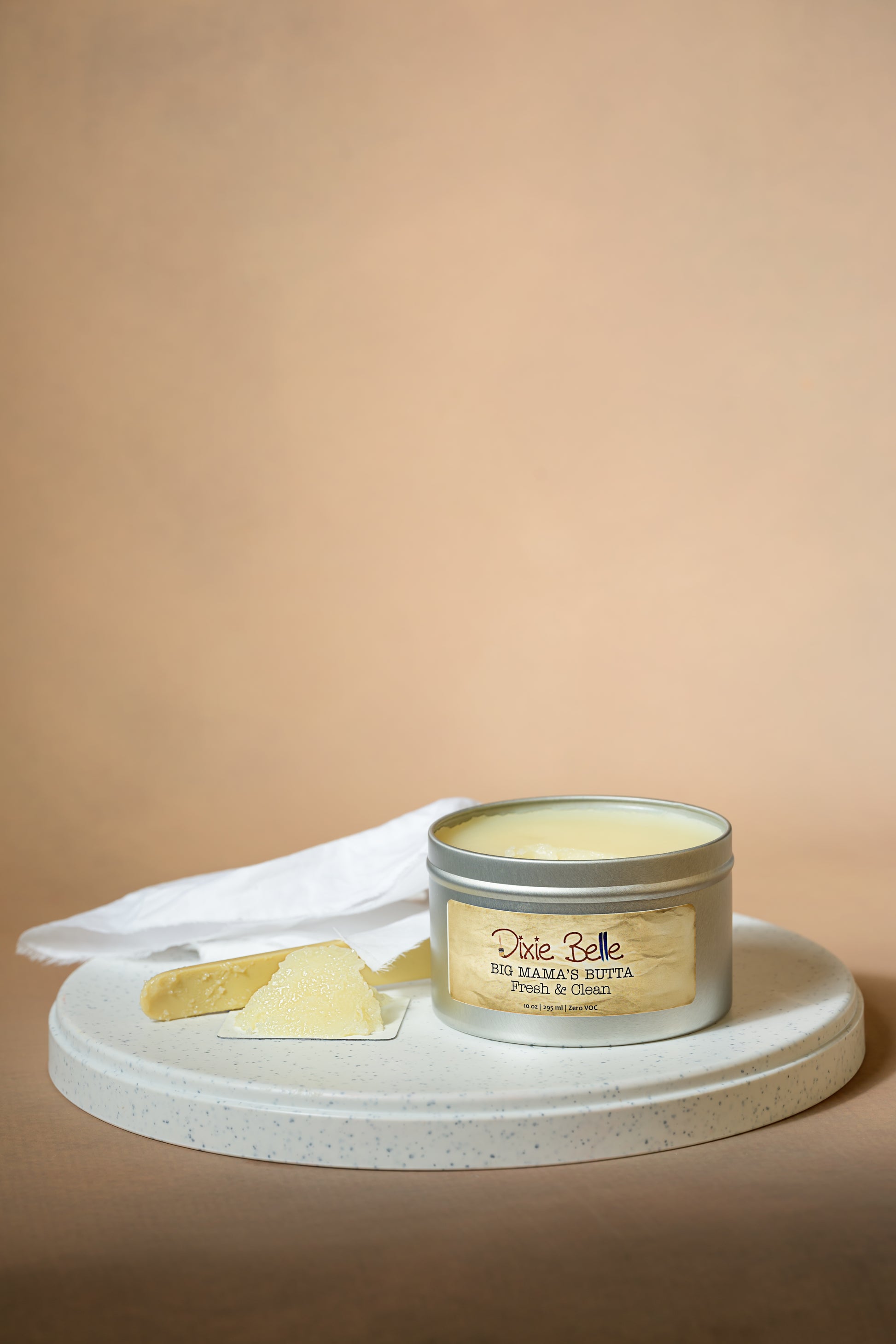 A tin labeled "Dixie Belle Big Mama’s Butta Ginger Zen" sits open on a round white tray, with a cloth and a small portion of the product scooped out beside it, against a neutral beige background.