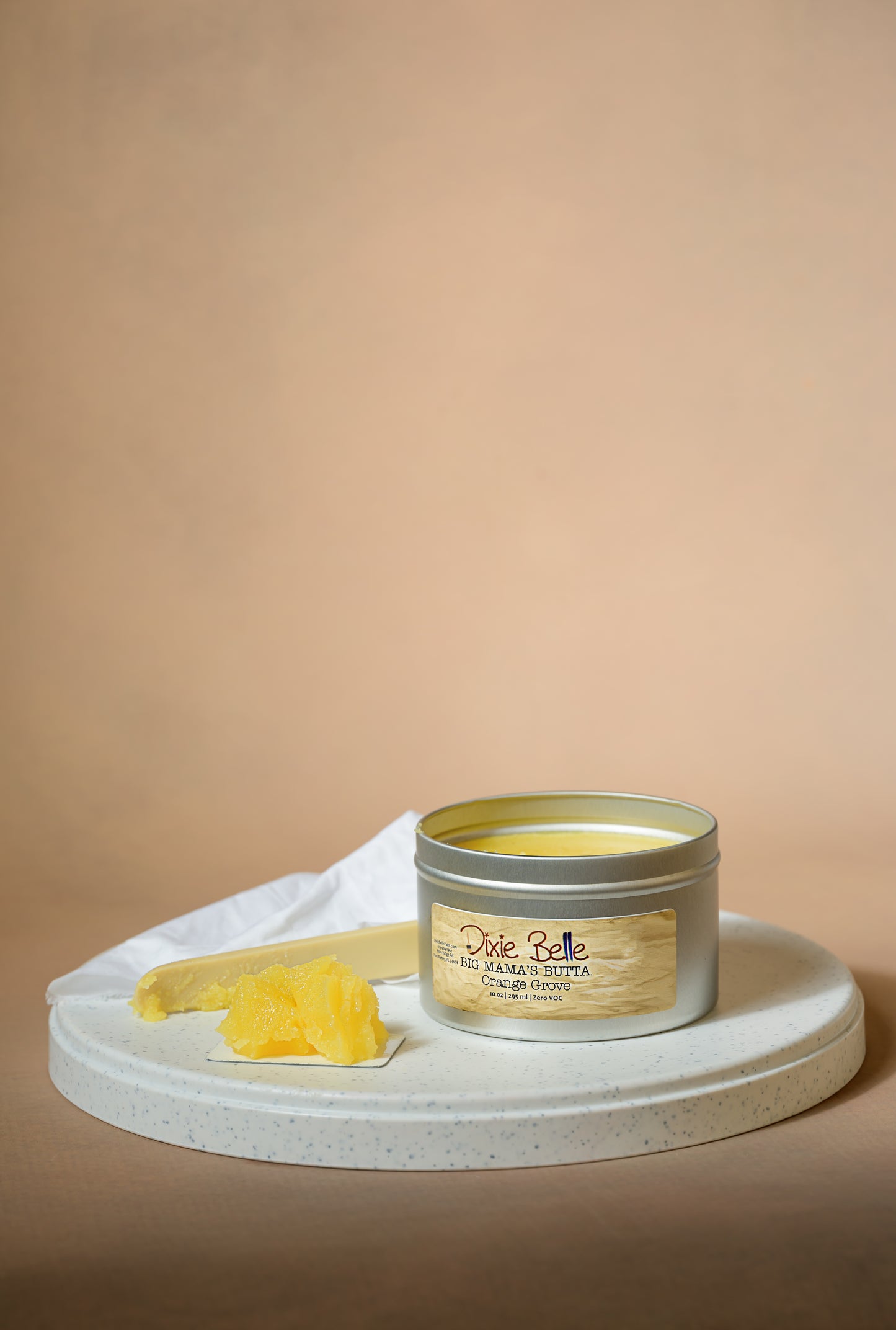 A tin of Dixie Belle Big Mama’s Butta Orange Grove sits open on a round white tray, with a scoop of yellow butter-like product next to it and a white cloth in the background, all set against a beige backdrop.