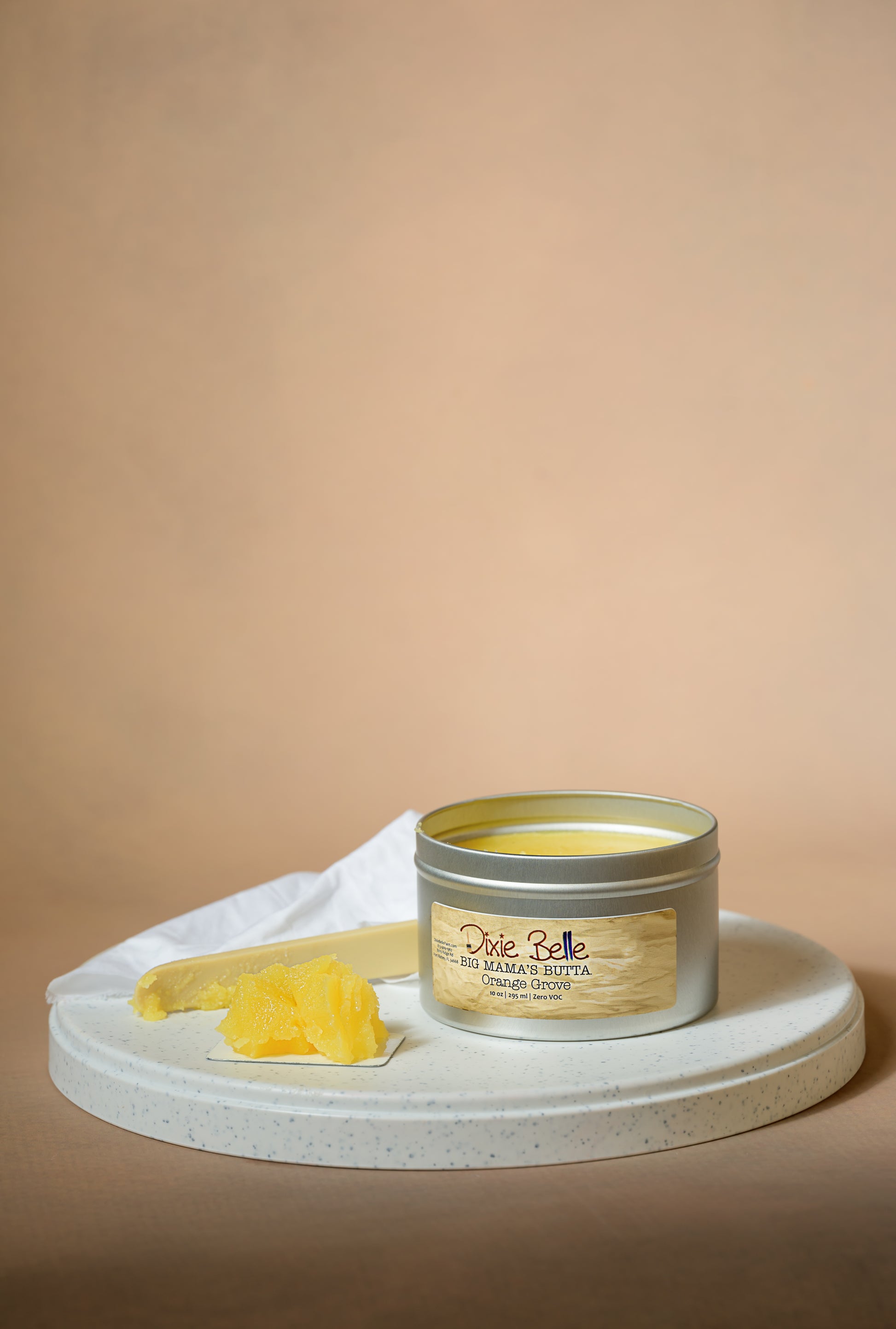 A tin of Dixie Belle Big Mama’s Butta Orange Grove sits open on a round white tray, with a scoop of yellow butter-like product next to it and a white cloth in the background, all set against a beige backdrop.