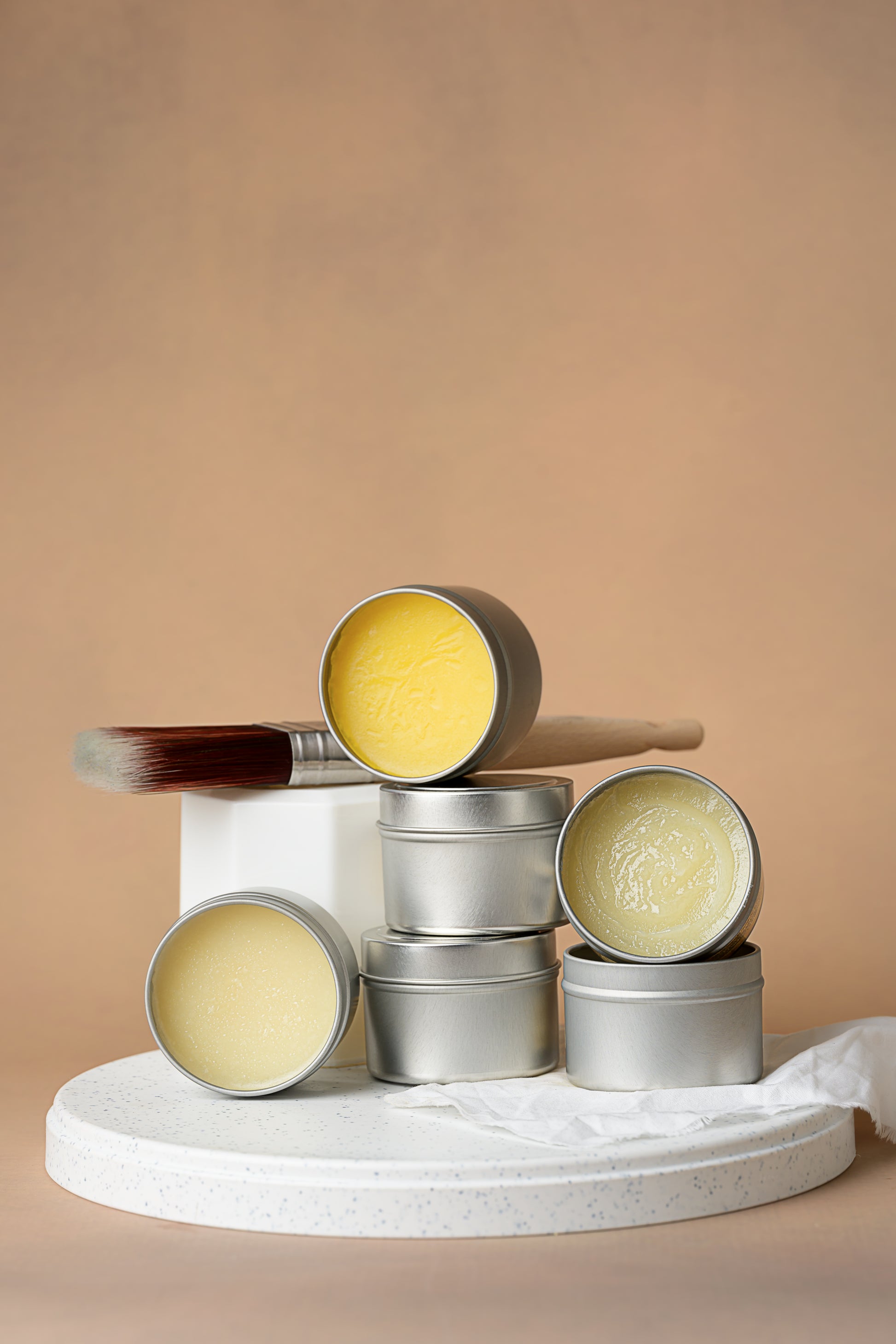 A stack of small silver tins, some open to show yellow and cream-colored balm, sits on a white round platform. A paint brush and a crumpled cloth are in the background against a beige backdrop.