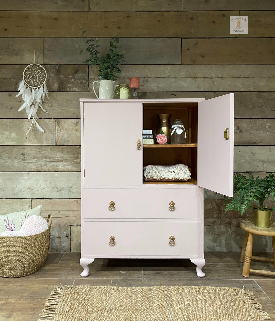 a pink cabinet painted in all in one paint with top right door open revealing a blanket and some books and vases with a pink flower.  On top is a pitcher with greenery and two candles sit next to that.  on wall is a dream catcher, to the left of cabinet is a wicker basket with pillows and to the right is a chair with a plant
