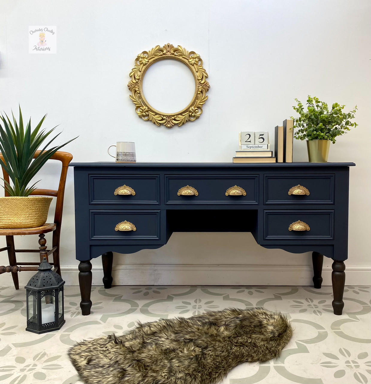A side table painted with all in one paint in a dark blue with gold pulls for hardware, staged with a plant in a wicker basket to the left, gold mirror on wall and plant and books with a coffee mug on top