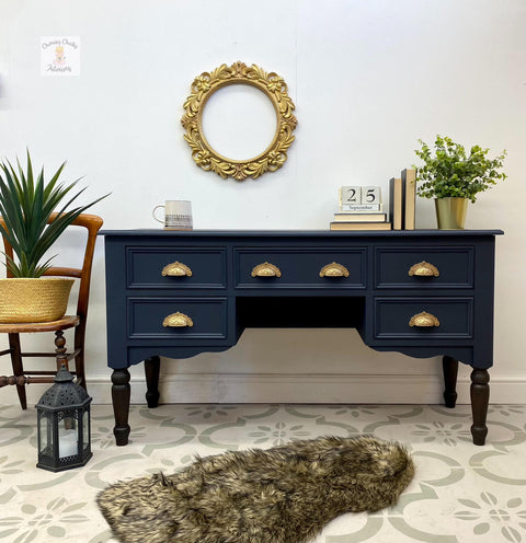 A side table painted with all in one paint in a dark blue with gold pulls for hardware, staged with a plant in a wicker basket to the left, gold mirror on wall and plant and books with a coffee mug on top
