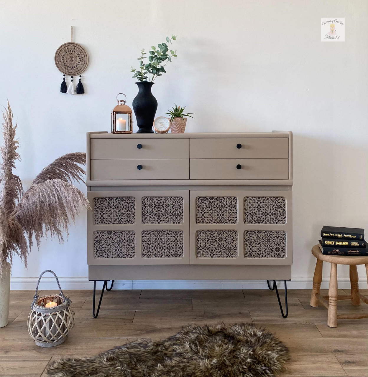 side table painted in a tan all in one paint, with four drawers on top and bottom has 8 squares that are inlays with decoupage. to the left is a container with pampas grass, on top is a candle and vases with plants, to the right is a stool with books and in front is a furry rug and a basket with a candle
