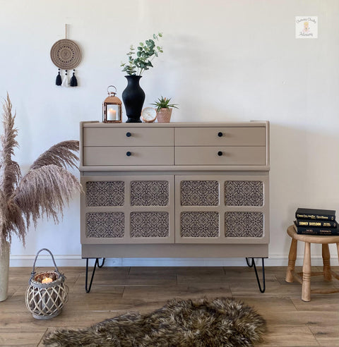 side table painted in a tan all in one paint, with four drawers on top and bottom has 8 squares that are inlays with decoupage. to the left is a container with pampas grass, on top is a candle and vases with plants, to the right is a stool with books and in front is a furry rug and a basket with a candle