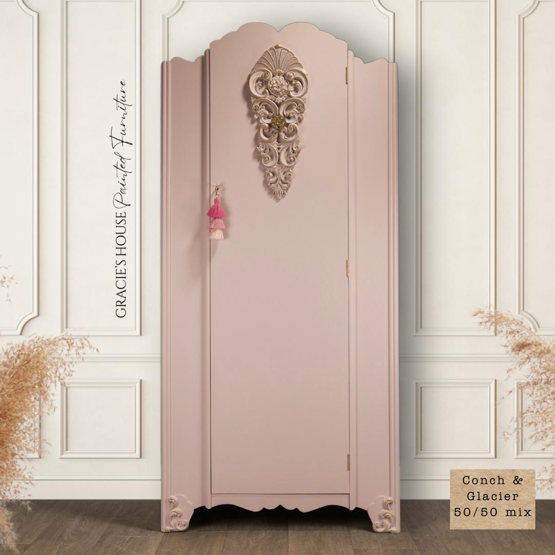 Conch Silk All-In-One paint is perfect for painting kitchen cabinets, bathroom cabinets and painting furniture
A vintage-style wardrobe painted in a soft pink shade, with ornate floral carvings at the top center and corners. Small tassels hang from the knob. Decorative text on the left reads “Gracie’s House Painted Furniture.” A beige swatch reads “Conch & Glacier 50/50 mix.”.
