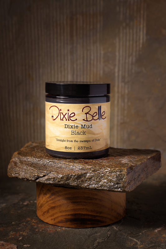 A container of Dixie Belle Dixie Mud Black sits open on a stone slab, with some paste coming out of the top and a small black spatula beside it. The background is neutral and textured.