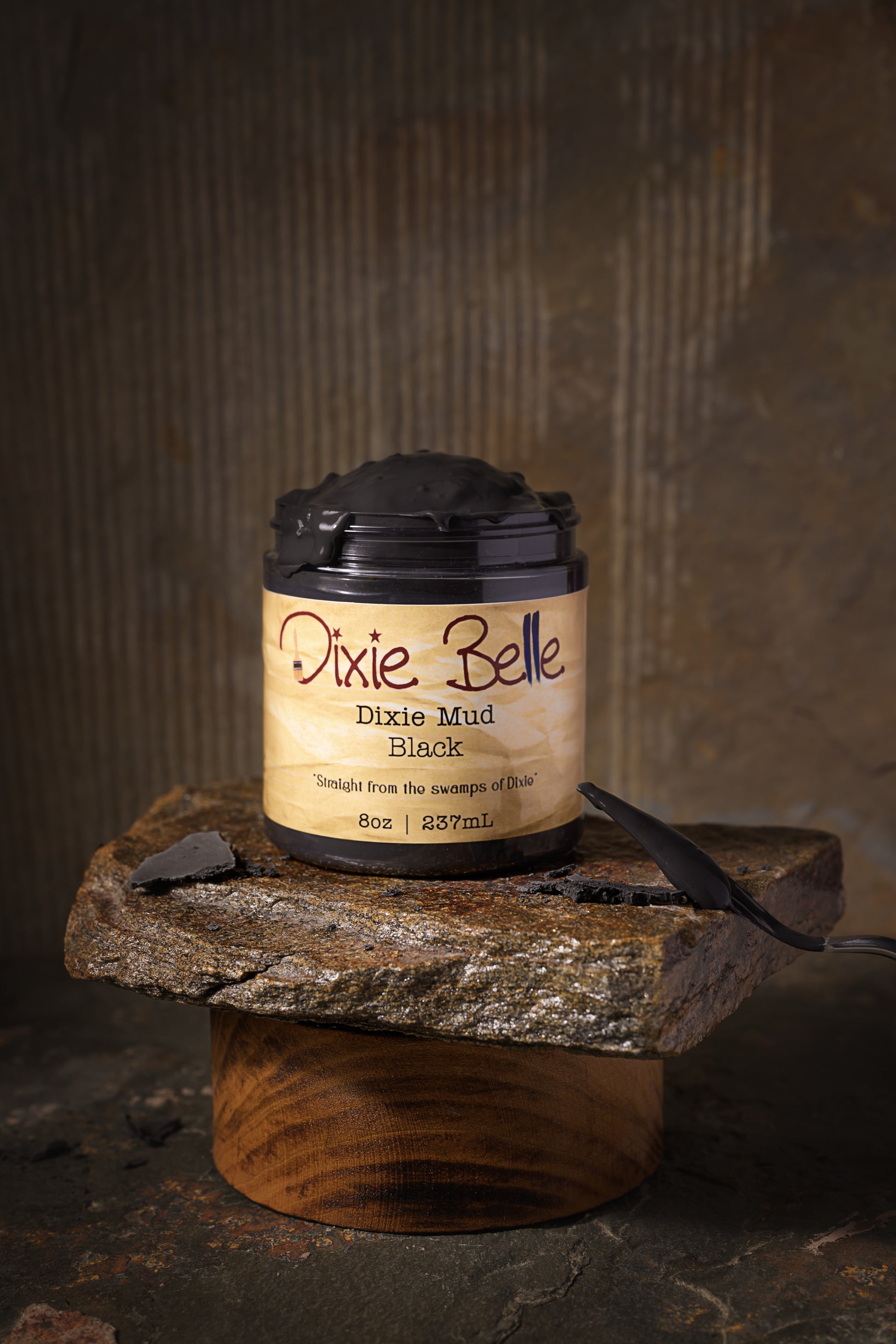 A jar of Dixie Belle Dixie Mud Black, 8 oz (237 mL), sits on a rough stone slab, which is supported by a round wooden base, against a rustic brown background.