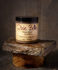 An open jar labeled "Dixie Belle Dixie Mud Brown" sits on a stone slab, with a small metal spatula holding some of the product in front. The background is rustic and earthy-toned.