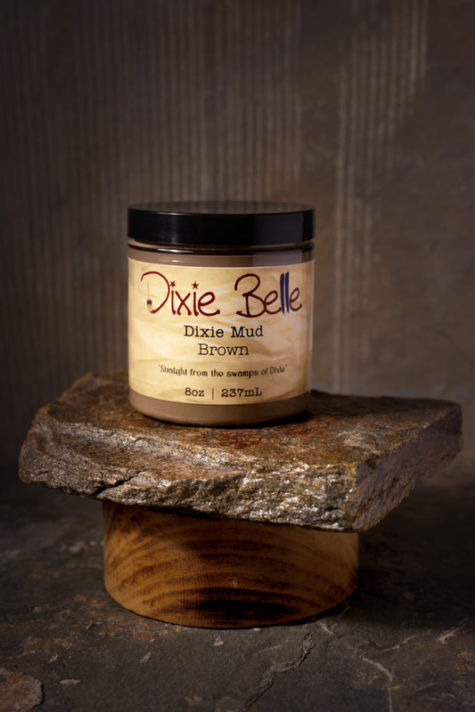 An open jar labeled "Dixie Belle Dixie Mud Brown" sits on a stone slab, with a small metal spatula holding some of the product in front. The background is rustic and earthy-toned.