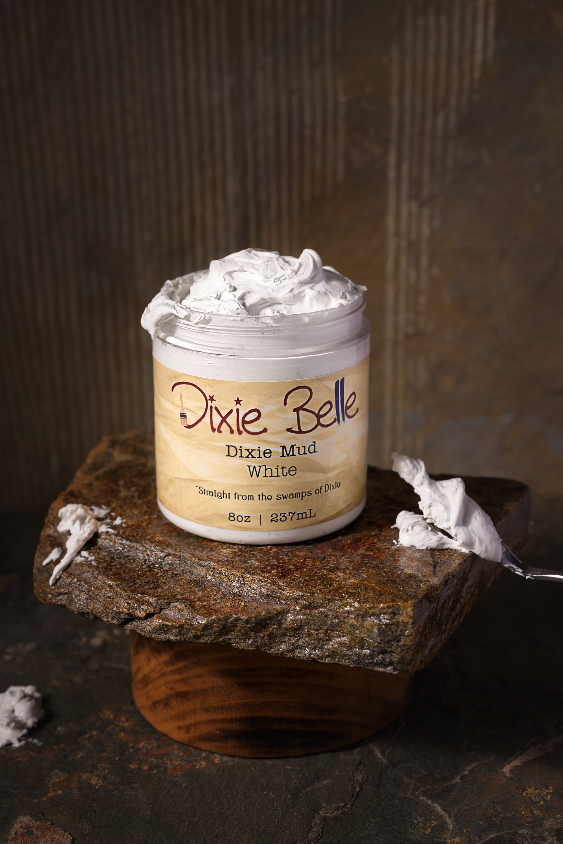 A jar of Dixie Belle Dixie Mud White, 8 oz (237 mL), sits on a stone slab with a wood base. The label reads "Straight from the swamps of Dixie." The background is brown and textured.
