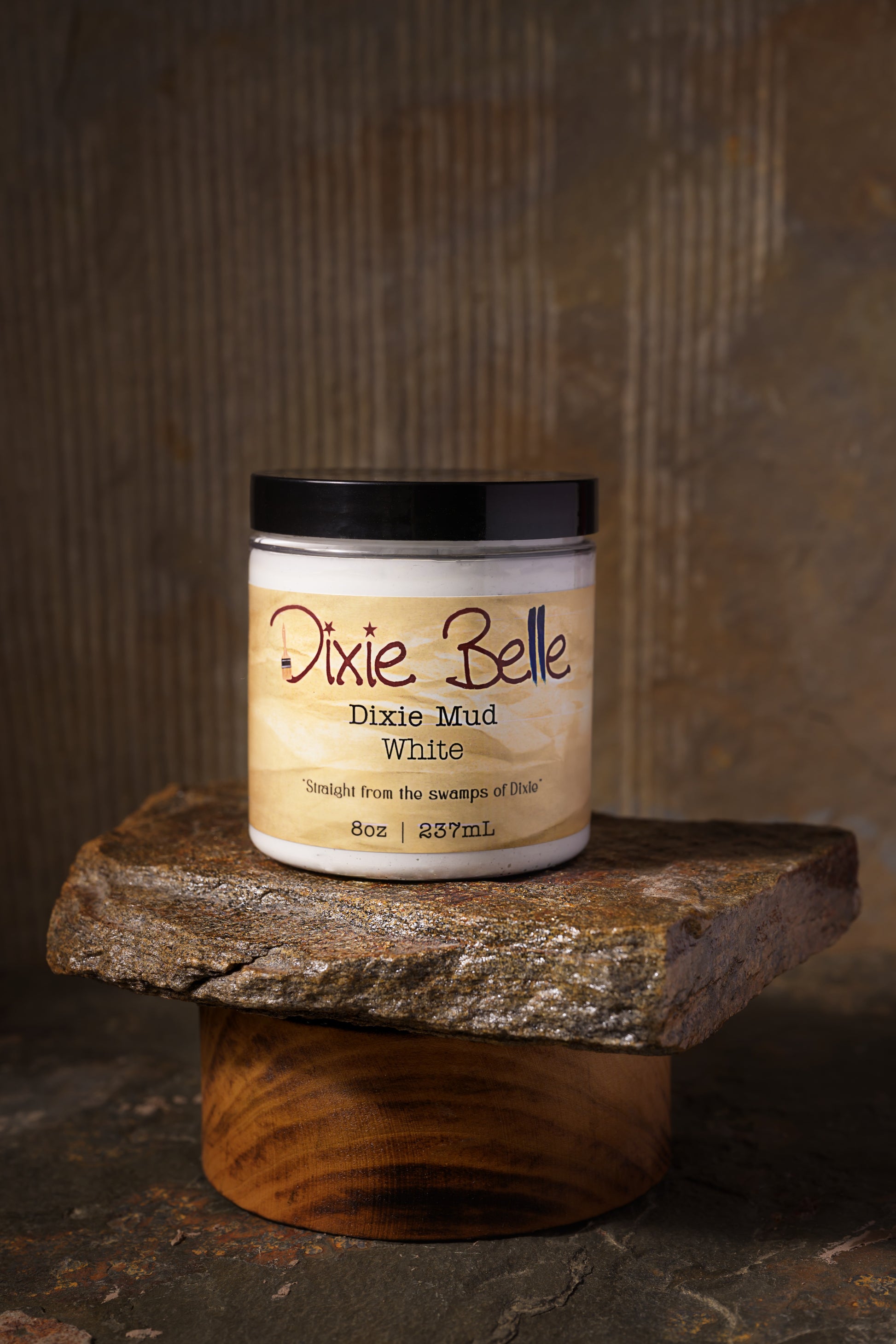 A jar of Dixie Belle Dixie Mud White sits on a stone slab. The jar is open, showing white mud paste with some spread on the stone and a tool. The label reads “Straight from the swamps of Dixie.”.
