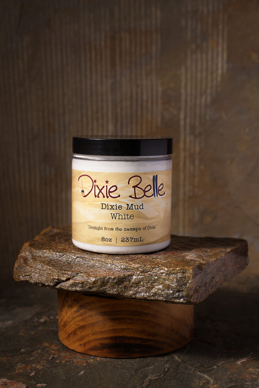 A jar of Dixie Belle Dixie Mud White sits on a stone slab. The jar is open, showing white mud paste with some spread on the stone and a tool. The label reads “Straight from the swamps of Dixie.”.