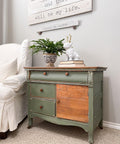 A side table painted with chalk paint in green color with the raw wood on one of the doors. a blue and white vase with a plant on top and a bulldog with antlers sitting on some books.  All this by a white fabric chair and a sign above it with a quote from Elsie De Wolfe