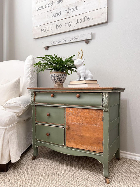 A side table painted with chalk paint in green color with the raw wood on one of the doors. a blue and white vase with a plant on top and a bulldog with antlers sitting on some books.  All this by a white fabric chair and a sign above it with a quote from Elsie De Wolfe