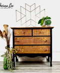 four drawer cabinet that the trim is painted with a dark black all in one paint and the drawers are natural wood.  To the left is a glass vase with dried plants, on top is staged with greenery and the wall has a gold modern geometric design