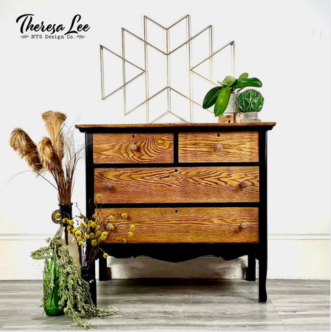four drawer cabinet that the trim is painted with a dark black all in one paint and the drawers are natural wood.  To the left is a glass vase with dried plants, on top is staged with greenery and the wall has a gold modern geometric design
