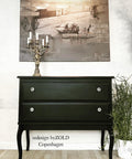 side table with two drawers, painted in a dark black all in one paint, staged with a candelabra on top and a picture on the wall of an old time city with truck and people.
