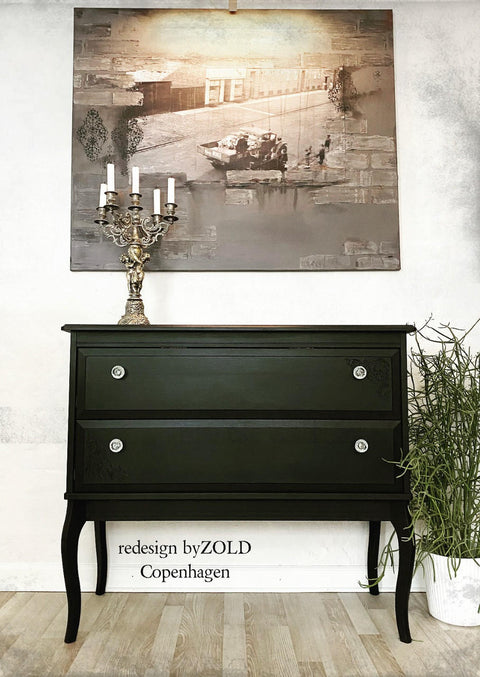 side table with two drawers, painted in a dark black all in one paint, staged with a candelabra on top and a picture on the wall of an old time city with truck and people.