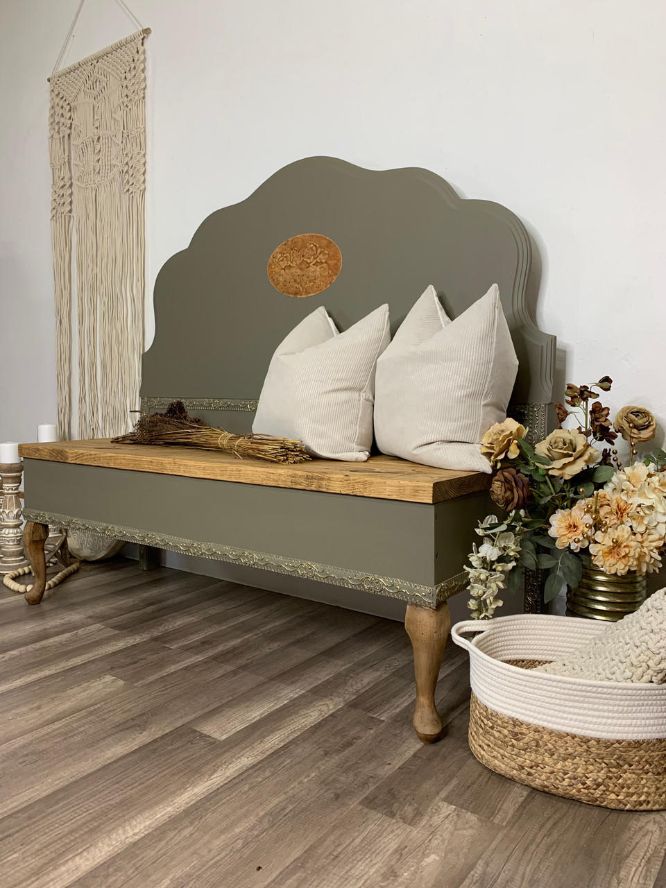 a long bench that is painted in all in one paint in a forest green color, two white pillows and a bundle of straw are staged on the bench. to the left of the bench is a wall handing made of rope and lots of plants in containers to the right of the bench.  Also a basket with a rug to the right.