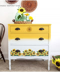 A bright yellow and white chalk painted three drawer dresser, the yellow is at the top and fades into the white.  At the bottom of the dresser is some sunflower rub on furniture transfer.  staged with green vases and some sunflowers.  to the left of the dresser is a brown chair and on the wall some wicker baskets.  front right is a basket with a sunflower.