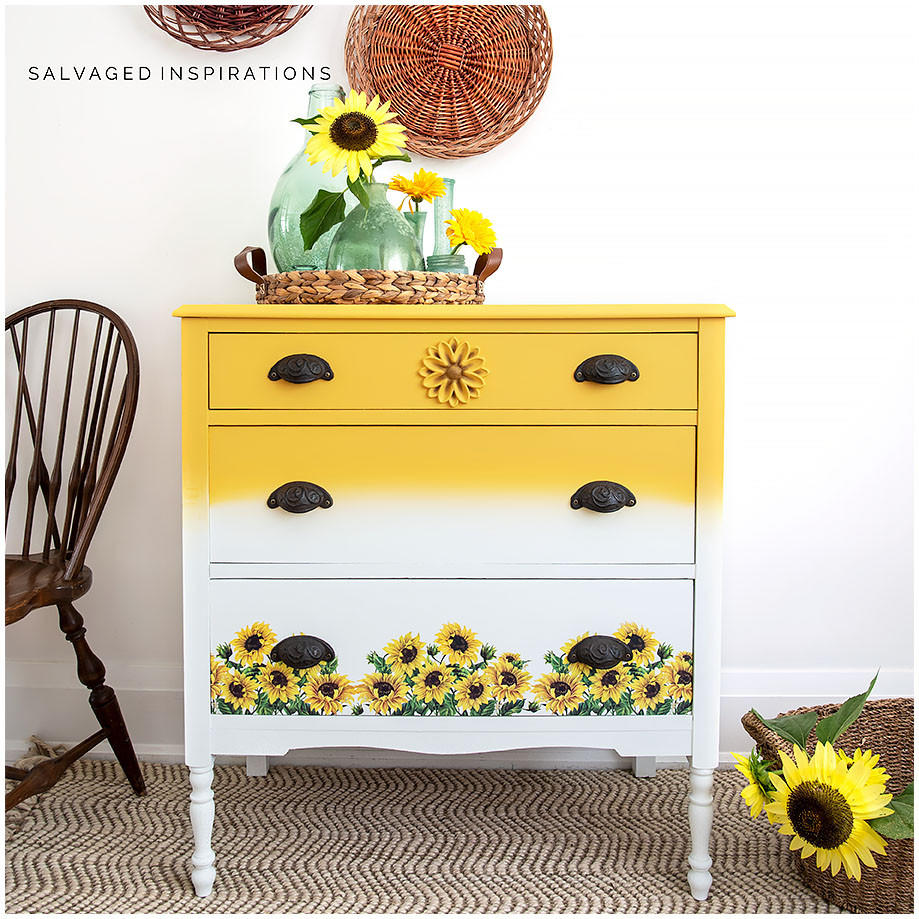 A bright yellow and white chalk painted three drawer dresser, the yellow is at the top and fades into the white.  At the bottom of the dresser is some sunflower rub on furniture transfer.  staged with green vases and some sunflowers.  to the left of the dresser is a brown chair and on the wall some wicker baskets.  front right is a basket with a sunflower.