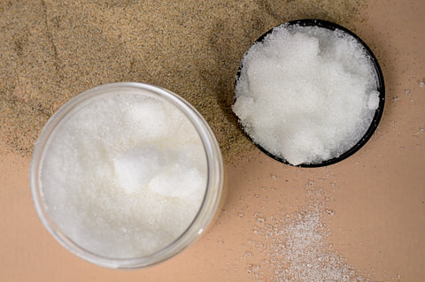 Top-down view of a jar and a black lid, both filled with whitish clear granules, placed on a beige surface sprinkled with sand and white granules.