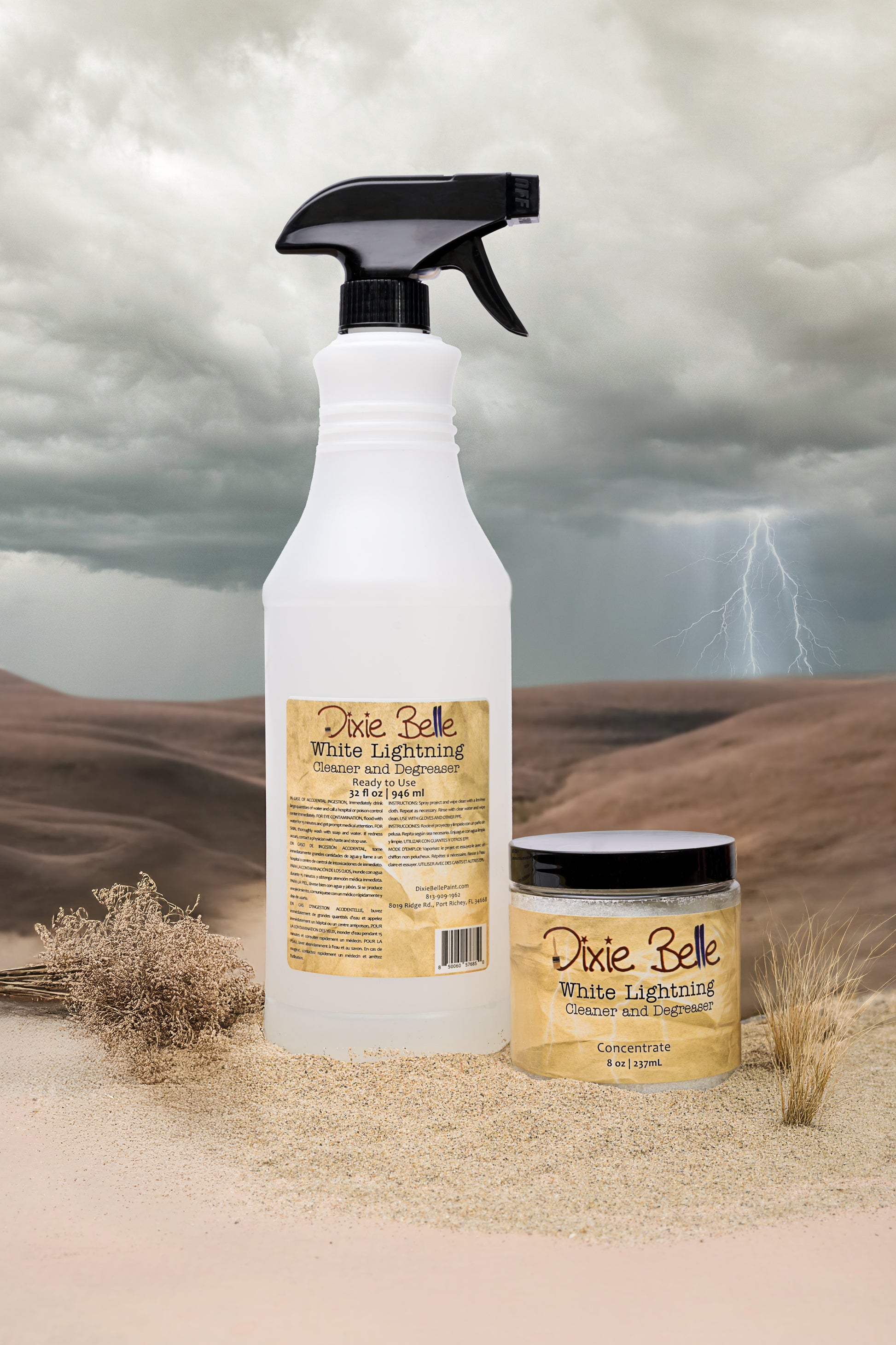 A spray bottle and a small jar labeled “Dixie Belle White Lightning Cleaner and Degreaser” sit on sand with dry plants, against a desert backdrop with dark clouds and a lightning bolt in the background.