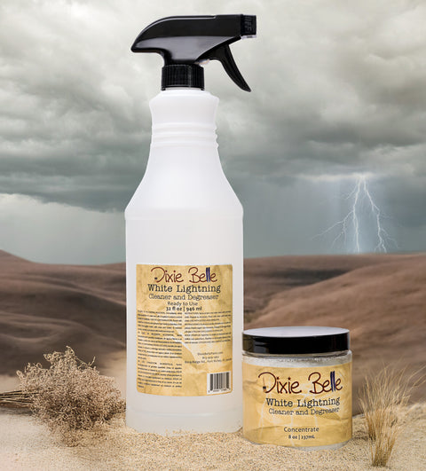A spray bottle and a small jar labeled “Dixie Belle White Lightning Cleaner and Degreaser” sit on sand with dry plants, against a desert backdrop with dark clouds and a lightning bolt in the background.