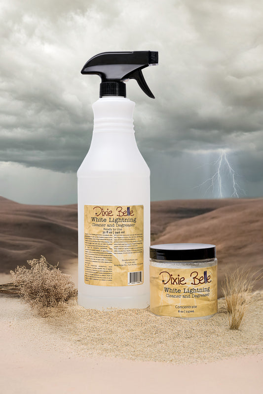 A spray bottle and a small jar labeled “Dixie Belle White Lightning Cleaner and Degreaser” sit on sand with dry plants, against a desert backdrop with dark clouds and a lightning bolt in the background.