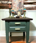 Dark green painted nightstand makeover with a dark wood top. Staged with 3 candlesticks, books and a picture frame on top, and a painting of a mermaid hanging on the wall behind the piece.