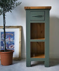 Dark green painted end table makeover, with two open cubbies and the top left as wood. Staged next to a potted tree and an old picture in a frame.