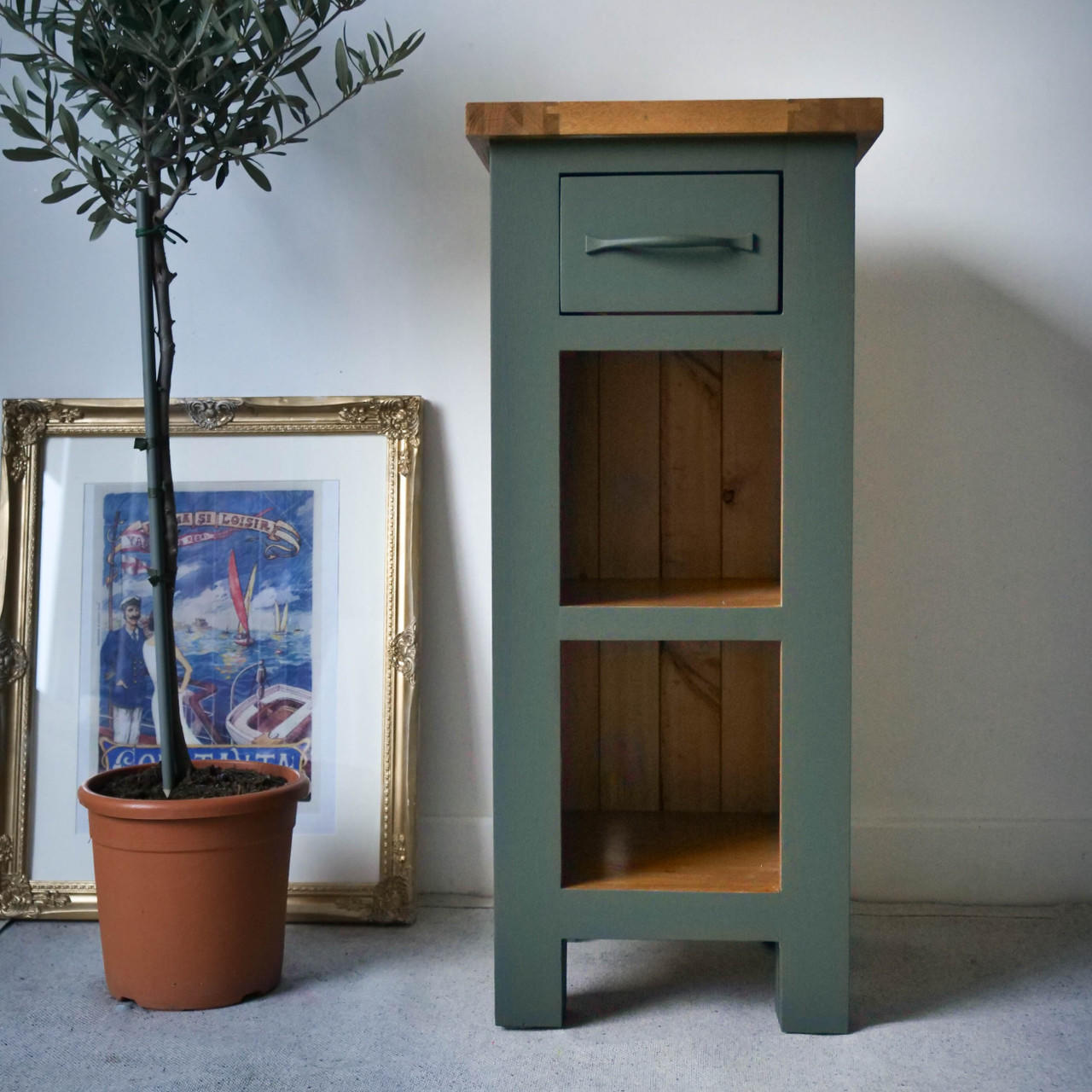 Dark green painted end table makeover, with two open cubbies and the top left as wood. Staged next to a potted tree and an old picture in a frame.