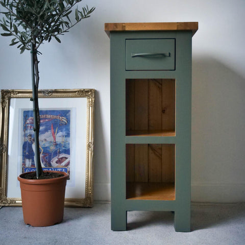 Dark green painted end table makeover, with two open cubbies and the top left as wood. Staged next to a potted tree and an old picture in a frame.