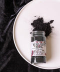 A jar labeled "Velvet Touch Black" lies on a white plate, with some black powder spilled beside it. The plate is set on a black velvet surface.
