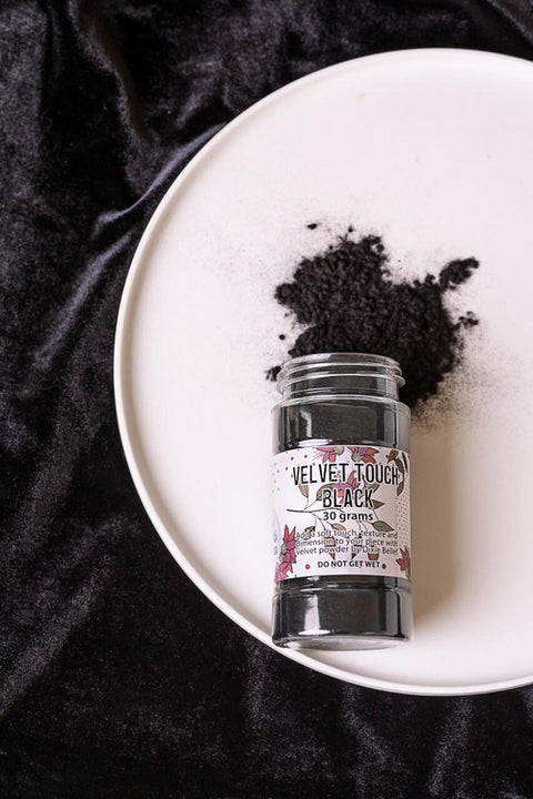 A jar labeled "Velvet Touch Black" lies on a white plate, with some black powder spilled beside it. The plate is set on a black velvet surface.